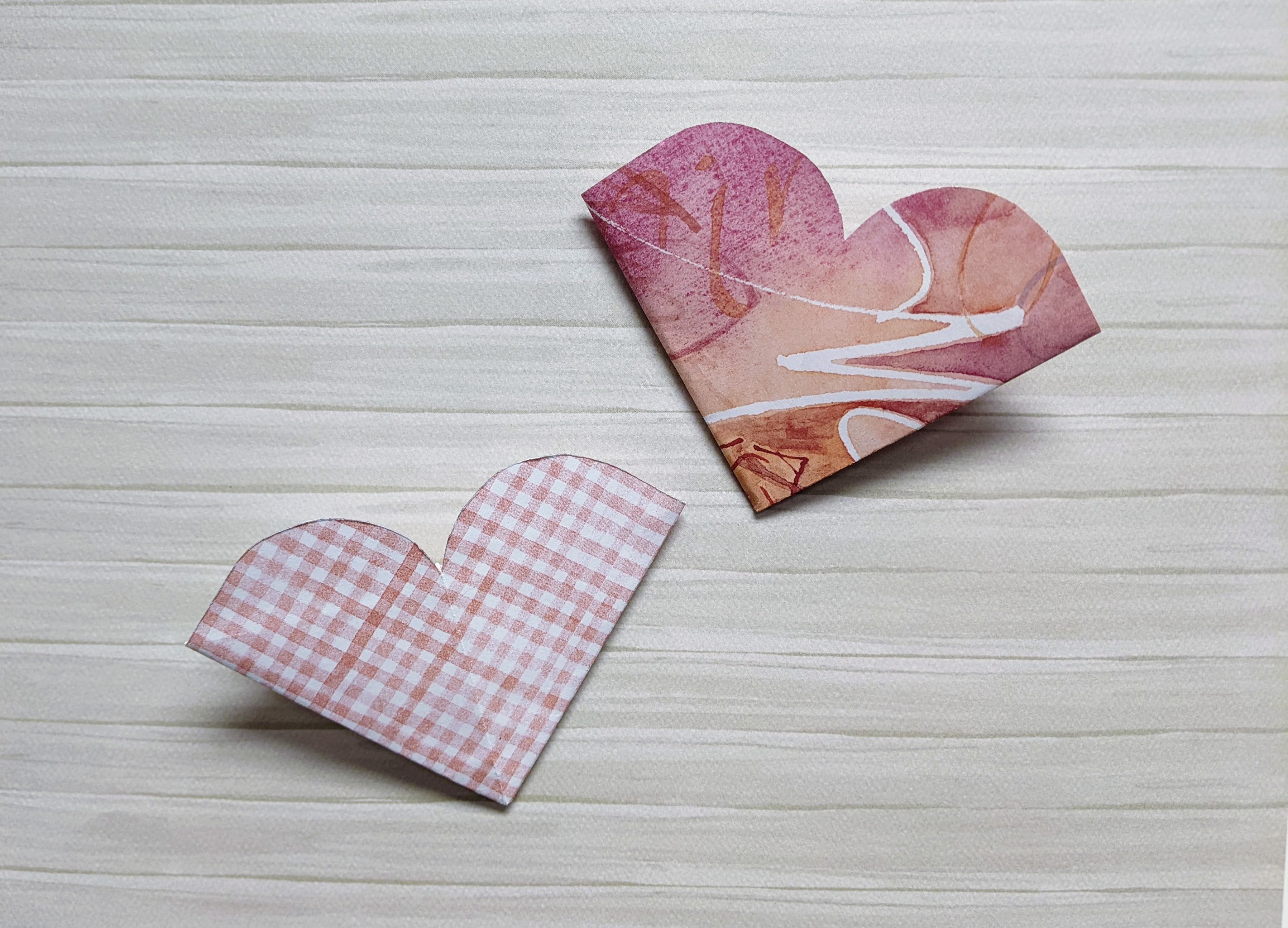 Two heart-shaped bookmarks. The one on the left is a white and pink gingham design, and the one on the right is an abstract design with pinks, white, and orange.