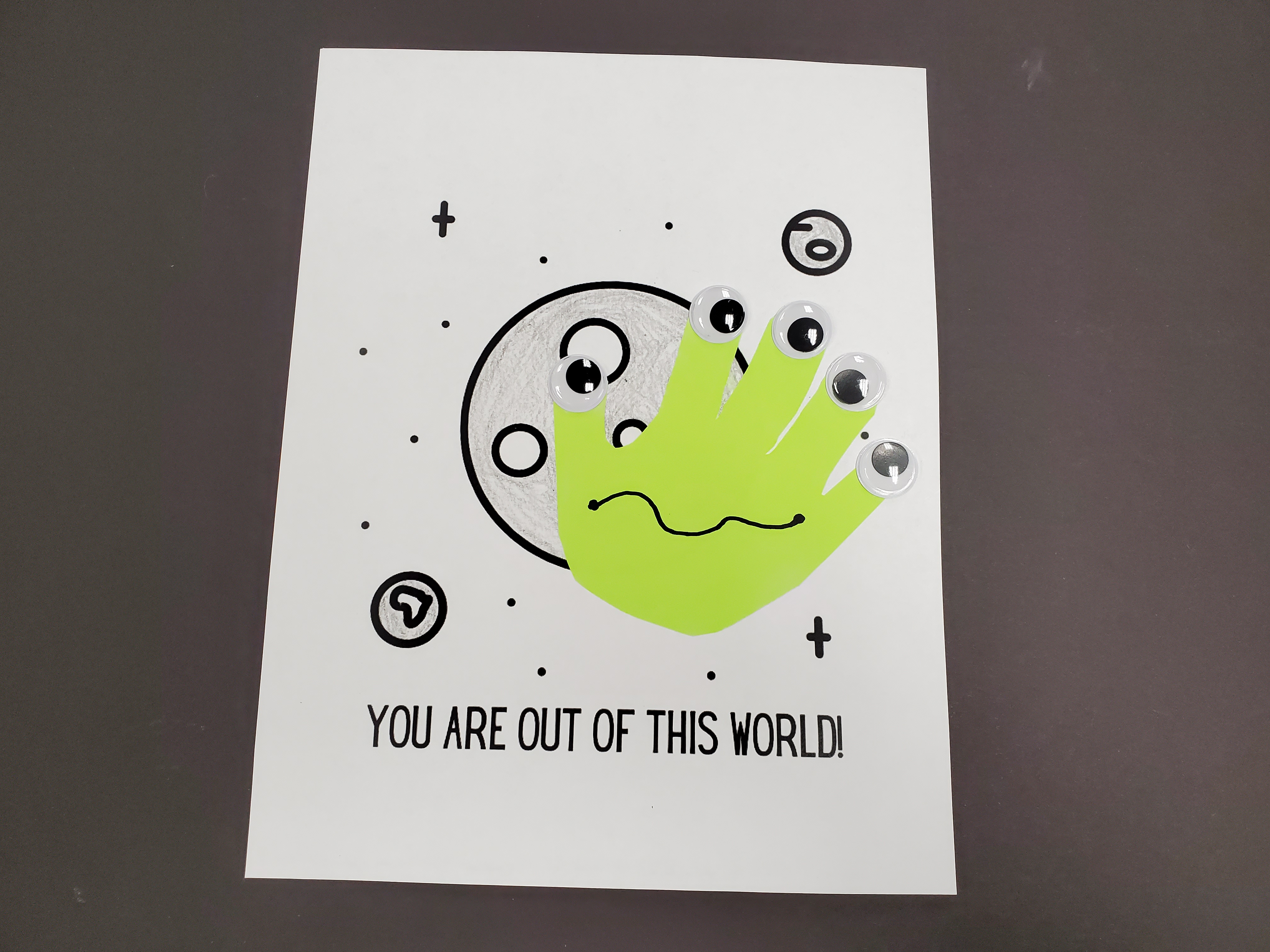Image of a bright green handprint cutout glued to a white sheet of paper with an image of the moon. The white sheet of paper has the words "You are out of this world" printed on it. The green handprint cutout has five googly eyes, one at the tip of each finger, to give the appearance of an alien creature.