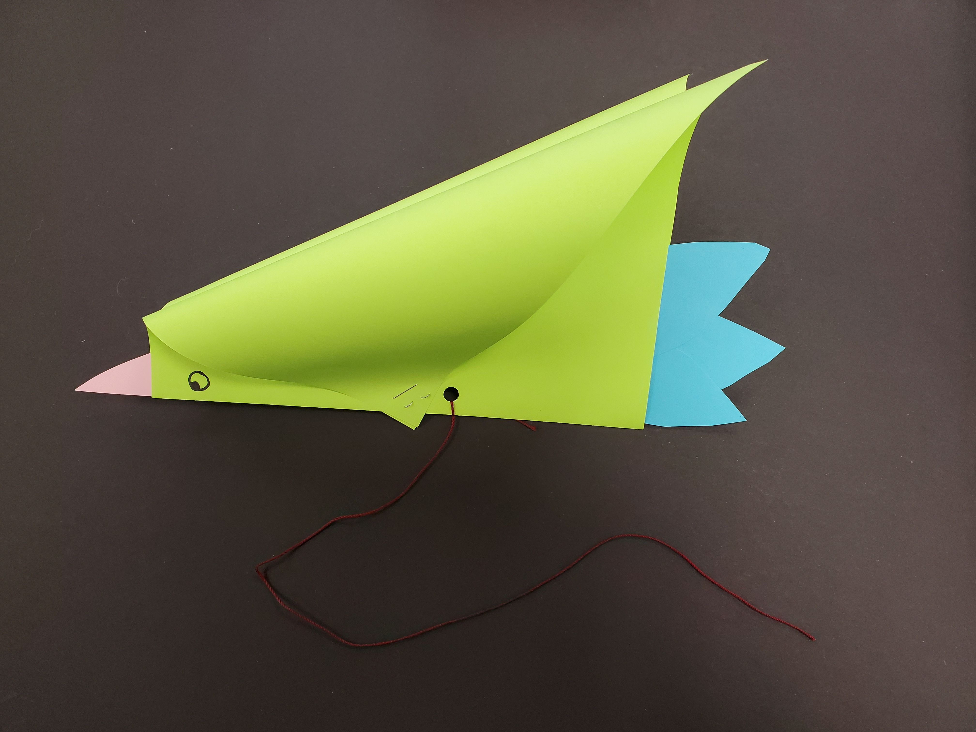 Image of a colored paper craft meant to resemble a bird. The body is made of bright green paper, the beak is cut from pink paper, and the three tail feathers are cut from blue paper. A red string is tied to the craft.