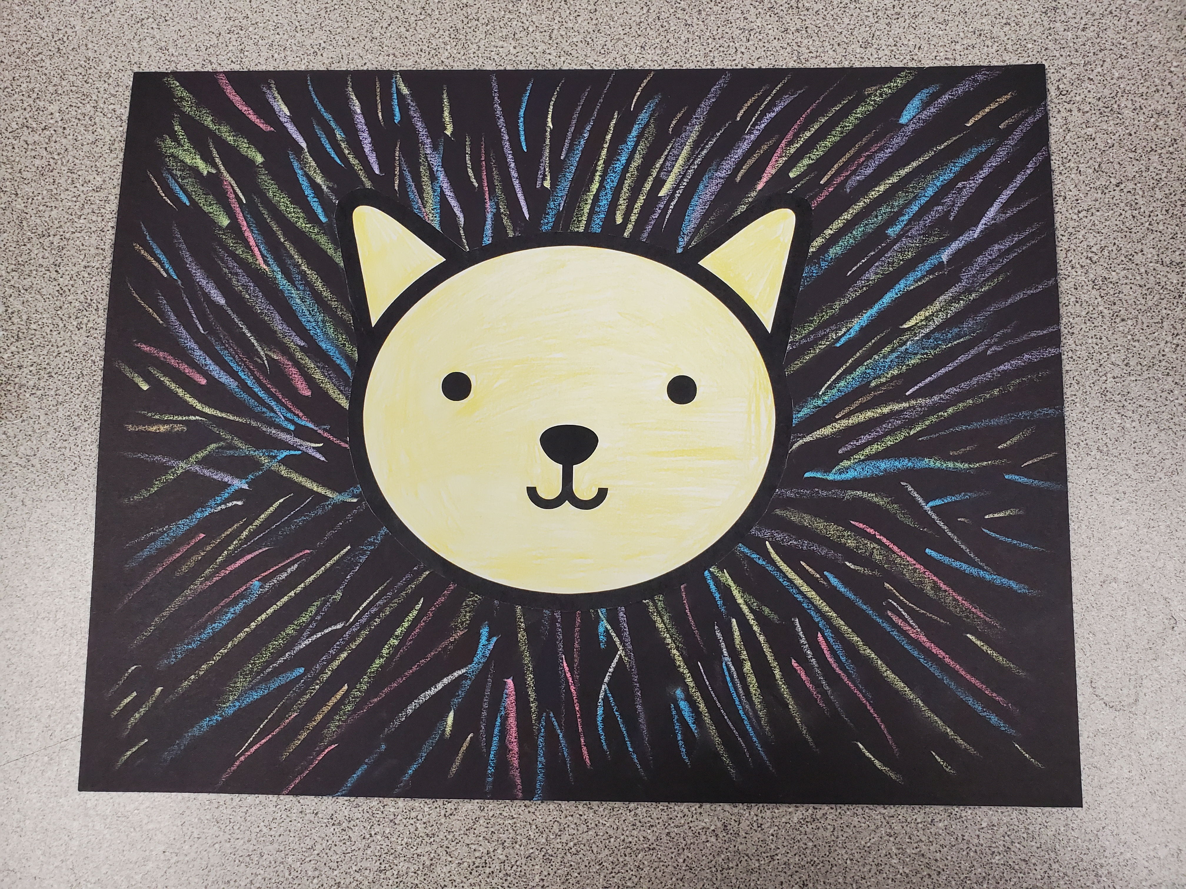 Image of a cartoon yellow lion face glued to a black piece of paper. The black paper has been colored with various colors of chalk, to give the appearance of a colorful lion mane.
