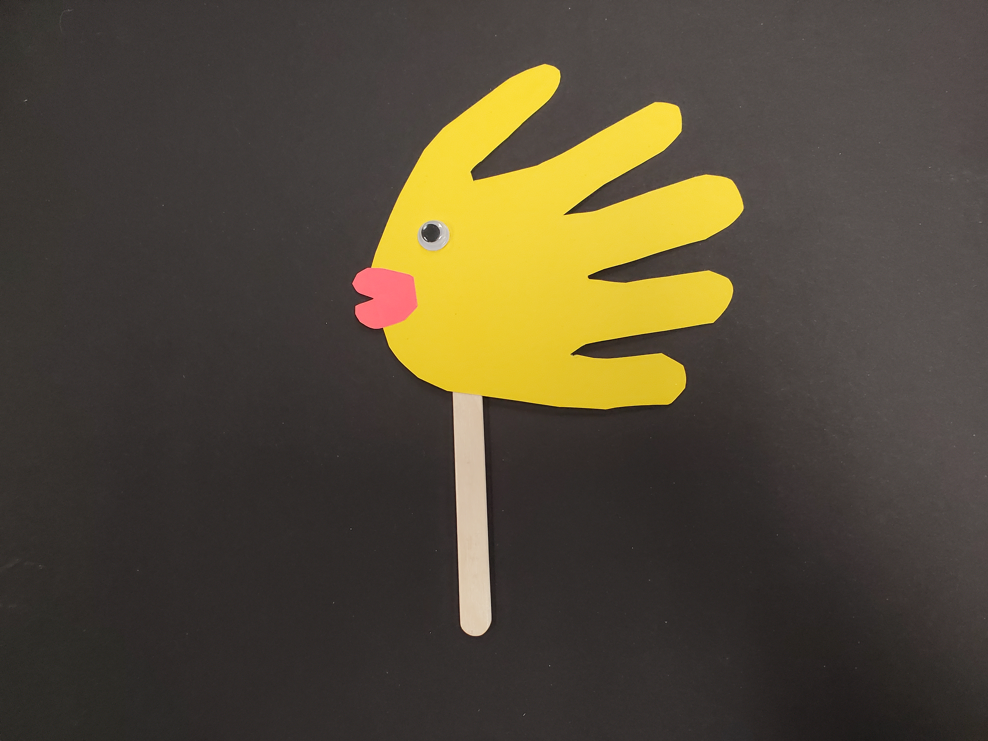 Image of a handprint fish puppet made from a cutout of a handprint on yellow paper, lips cut out of red paper and attached at the palm of the handprint, and a small googly eye sitting above the lips. All of this is attached to a popsicle stick so it can be used as a puppet.