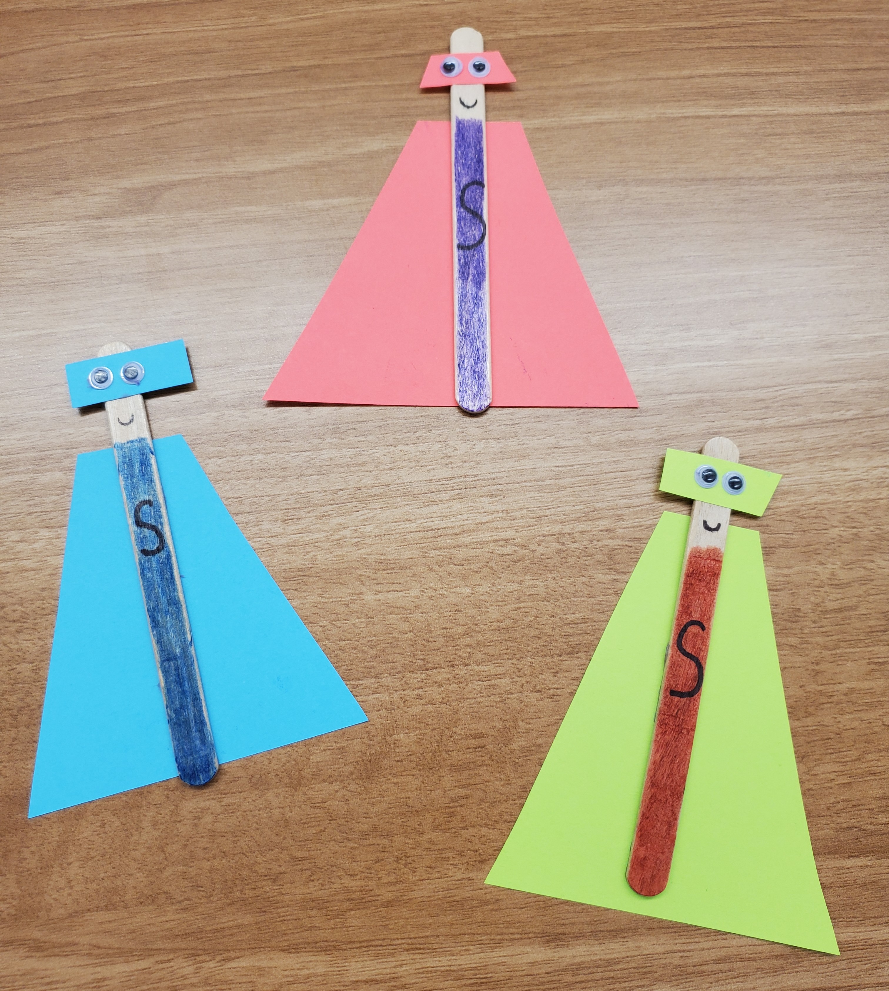 Example of three superhero popsicle stick crafts. Each craft has a different color cape and mask made from paper - one is blue, one is red, and one is yellow.