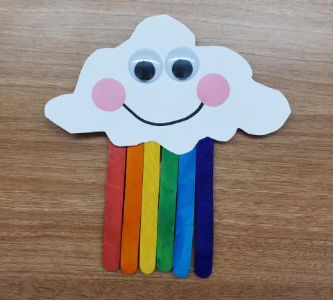 Photo of a row of rainbow popsicle sticks with a white cloud cutout attached. The cloud has googly eyes and a smiling face. 