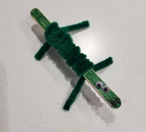 Photo of a craft of a crocodile made using a popsicle stick, green pipecleaners, and googly eyes.