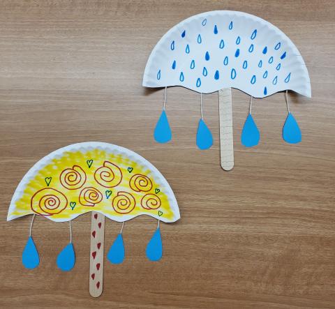 Photo of an umbrella craft made out of a half paper plate, popsicle stick, and blue construction paper. There are two crafts in the image, one has a white background with blue raindrops and the other has a yellow background with red swirls.